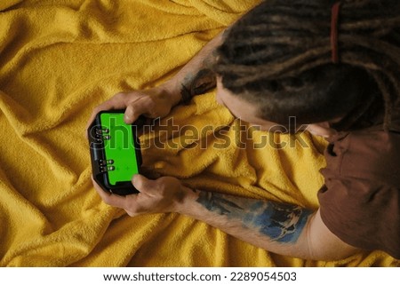 Young man lies inside on yellow blanket on bed and plays using phone and joystick, top view. Hands with multicolored tattoos close-up. Guy Uses Green Mock-up Screen Smartphone.