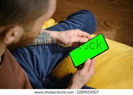 Guy Uses Green Mock-up Screen Smartphone. Young man sitting inside on yellow blanket on bed and uses phone to work or browse the Internet online, top view. Hands with multicolored tattoos close-up.