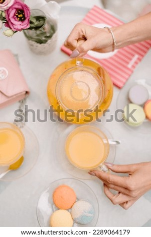 Cafe guests sit at a table and pour orange juice into glass cups. View from above. There are macaroons on the table in plates. There is a rose in a vase of water. Summer day