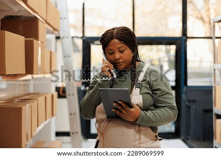 Stockroom employee talking at landline phone with client while checking online order on tablet computer. African american manager preparing packages for delivery in warehouse. Merchandise concept Royalty-Free Stock Photo #2289069599