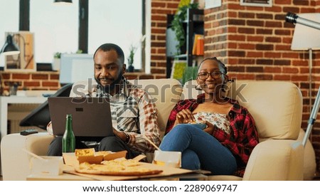 Cheerful couple watching television together at home, boyfriend browsing internet on laptop and girlfriend eating bowl of chips. Life partners enjoying action movie with beer bottles. Tripod shot. Royalty-Free Stock Photo #2289069747