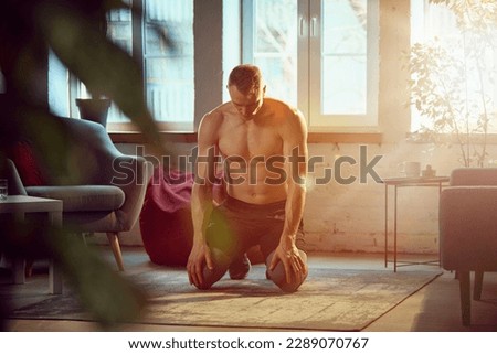 Young man with strong, muscular, relief body posing shirtless, doing home training on daytime with sunlight. Breathing. Concept of sportive lifestyle, body and health care, fitness, health Royalty-Free Stock Photo #2289070767