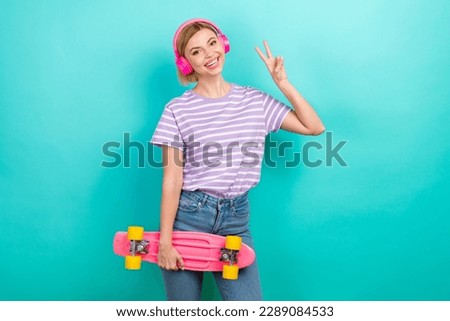 Photo of young youth girl wear t-shirt showing v-sign use air headphones hold longboard cheerful cadre isolated on cyan color background