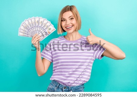 Photo of young promoter satisfied girl wear striped t-shirt thumb up like credit company hold dollars isolated on aquamarine color background