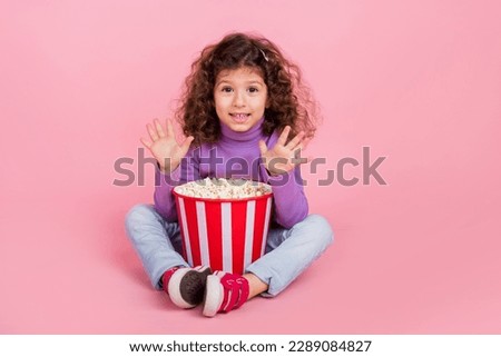 Full length photo of charming sweet little child dressed purple pullover waving arms hi eating pop corn isolated pink color background
