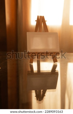 White canvas on wooden easel on a glass table vertical photo. School of drawing, creativity, creating pictures, works of art, masterpiece concept. Creative crisis. Warm lighting indoors in a room.