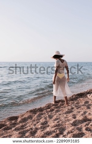 Vertical photo back view of a full-length woman walking on the beach in the summer in a beach lace knitted white dress and a yellow swimsuit