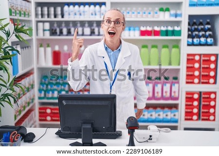 Young caucasian woman working at pharmacy drugstore pointing finger up with successful idea. exited and happy. number one. 