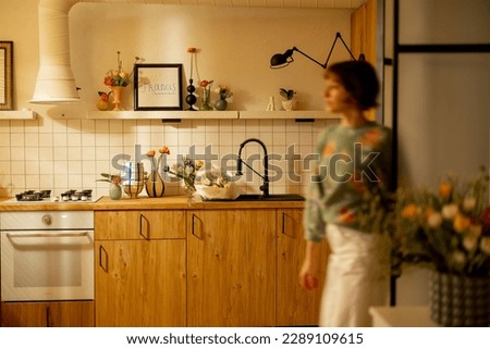 Modern kitchen interior made in natural materials with wooden facades and open shelves decorated with flowers. motion blurred female figure in front