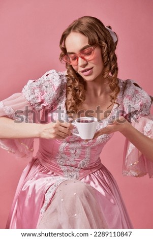 Morning drink. Portrait of happy charming princess wearing beautiful dress and sunglasses holding cup of coffee and looking on it over studio background. Concept of medieval, beauty, food, ad