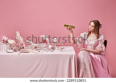 Food of the gods. Fairy tale blond queen, princess wearing renaissance dress holding big sandwich near the laid festive table on pink studio background. Concept of medieval, beauty, food, baroque, ad