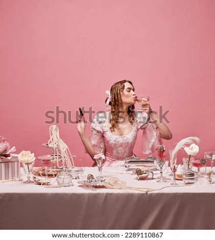 Happy birthday. Shot of adorable blond princess wearing fancy pink dress and sitting at beautifully laid table with wine glass and lipstick over studio background. Concept of beauty procedures