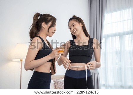 Asian female friends exercising at home. Young woman in workout clothes holding a glass of orange juice and measuring her waist with a tape measure