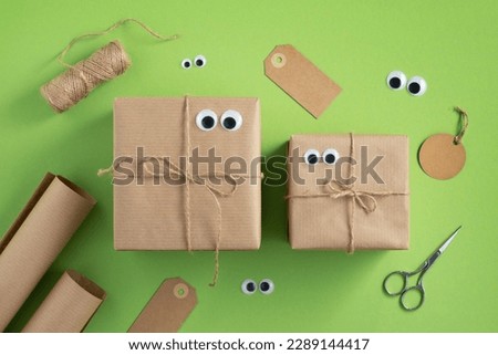 Funny gift boxes with eyes and wrapping materials. Jute rope, wrapping paper roll, scissors and gift tags on bright green background. Royalty-Free Stock Photo #2289144417