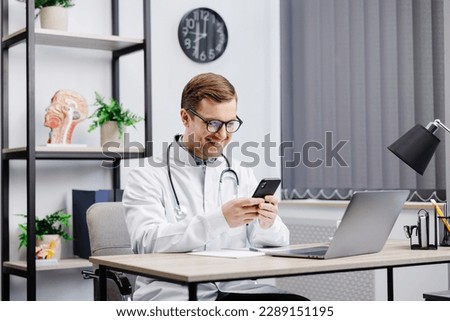 Male doctor medical worker in modern clinic wearing eyeglasses and white coat uniform using cell mobile smartphone apps, sitting at laptop computer. Medicine technologies health care concept