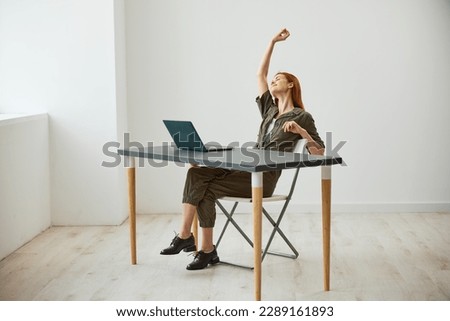 a very happy woman sits at a laptop and rejoices at the completed tasks raising her hands up