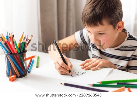 A child boy draws on white paper with colored pencils while sitting at a table