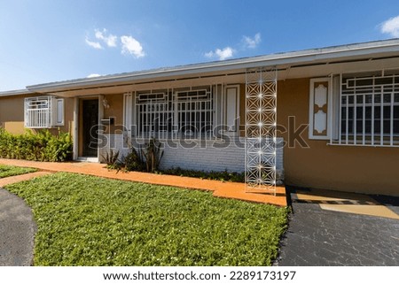 Traditional style suburban home located in the Westchester neighborhood of Miami-dade county, terracotta colored walls with large short grass park around it, privet fence, blue sky, covered patio