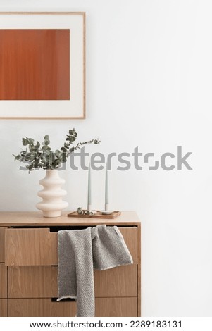 wall picture , candlesticks on wooden tray near modern vase with eucalyptus branches and towel hanging from open chest drawer in living room with elegant interior