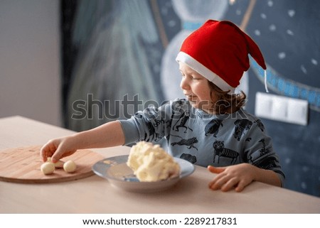 A blond long-haired European child in a santa hat in pajamas sits at a table and sculpts white homemade candies against a chalk wall
