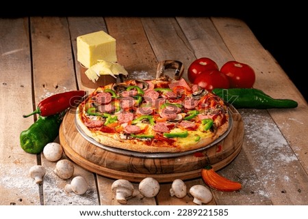 Pizza surrounded by peppers, tomatoes, mushrooms and a block of mozzarella cheese on a rustic wooden table against a black background