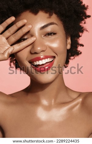 Vertical shot of gorgeous Black woman with healthy smile, touches her face and looks happy, has shiny glowing skin and body, spa after effect, has nourished and moisturized face, pink background. Royalty-Free Stock Photo #2289224435