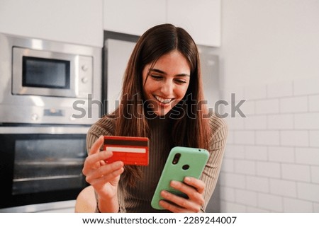 Young caucasian female with white perfect teeth smiling and using a cell phone app to purchase and pay with a credit card sitting at home kitchen. Woman spending money on online shopping on internet