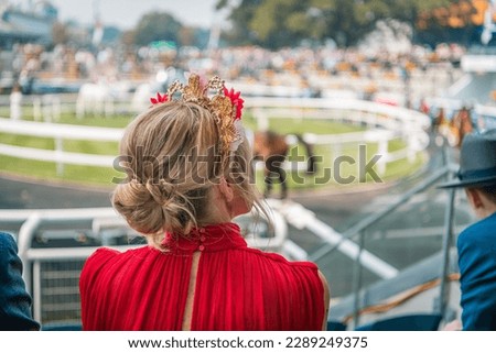 Woman in red dress wearing beautiful fascinator watching horses parading before the race Royalty-Free Stock Photo #2289249375