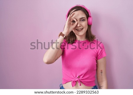 Blonde caucasian woman listening to music using headphones smiling happy doing ok sign with hand on eye looking through fingers 