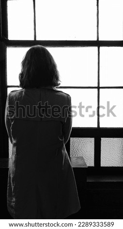 A young woman dressed in  retro-style trench coat stands on the windowsill with her back turned and looks out the window.