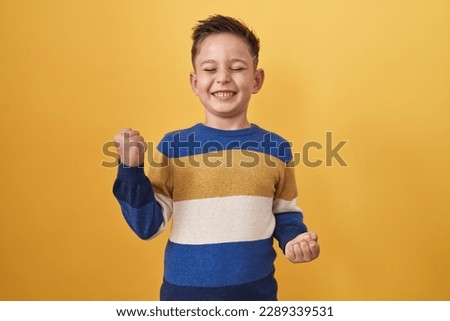 Little hispanic boy standing over yellow background celebrating surprised and amazed for success with arms raised and eyes closed  Royalty-Free Stock Photo #2289339531