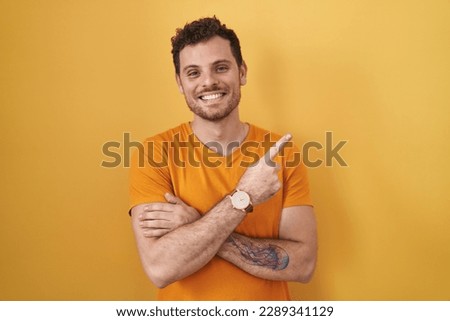 Young hispanic man standing over yellow background with a big smile on face, pointing with hand finger to the side looking at the camera. 