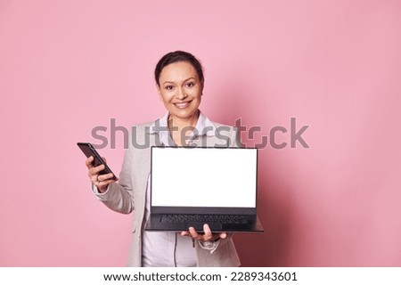 Multi-ethnic positive women, business person, entrepreneur, sales manager holding mobile phone, smiling looking confidently at camera, showing laptop with white blank digital screen on pink background