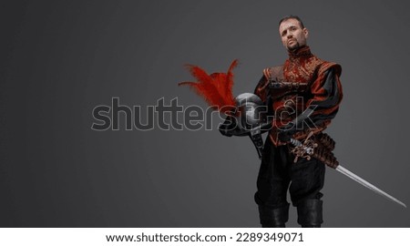 Portrait of fashionable musketeer swordsman with helmet isolated on gray.