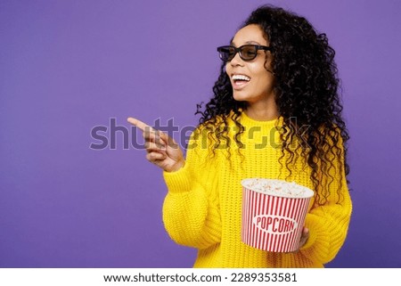 Young fun woman of African American ethnicity in 3d glasses wear casual yellow sweater watch movie film hold bucket of popcorn in cinema point finger aside isolated on plain purple background studio