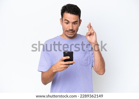 Young handsome man over isolated white background using mobile phone with fingers crossing