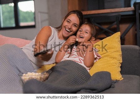 Happy young mom and child girl laughing holding snack popcorn remote control enjoy funny television comedy movie