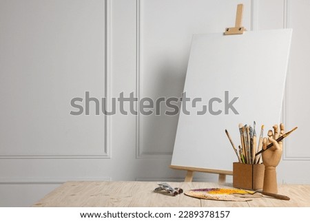 Easel with blank canvas, hand model and different art supplies on wooden table near white wall. Space for text