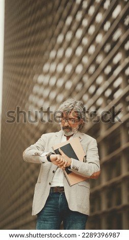 Mature businessman holding folder with business documents is standing outside on modern buildings background. Middle aged manager looks at his wristwatch