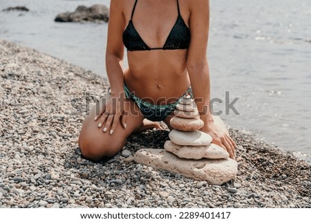 Woman bilds stones pyramid on seashore on a sunny day on the blue sea background. Happy holidays. Pebble beach, calm sea, travel destination. Concept of happy vacation on the sea, meditation, spa.