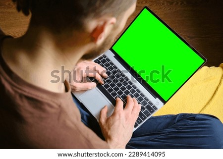 Young man sitting on yellow blanket on bed and uses laptop to work or browse Internet online, watching movie or TV series, top view. Copy space for advertising or design. Green Mock-up Screen.