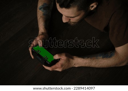 Young man lies inside on dark wooden floor and plays using mobile phone and joystick, close up top view. Guy Uses Green Mock-up Screen Smartphone.