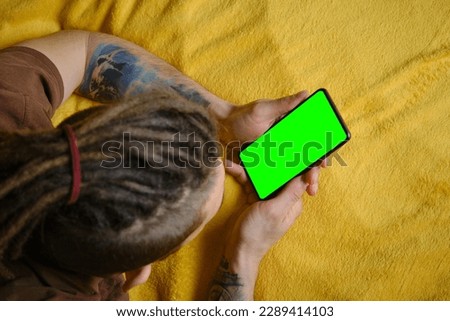 Guy Uses Green Screen Smartphone. Young man lies on yellow blanket and uses phone to work or browse the Internet online, watching movie or TV series, top view. Copy space for advertising and design.