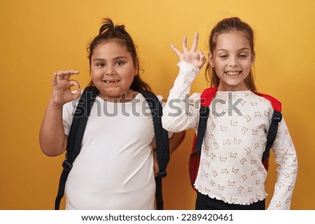 Two little girls friends wearing student backpack doing ok sign with fingers, smiling friendly gesturing excellent symbol 