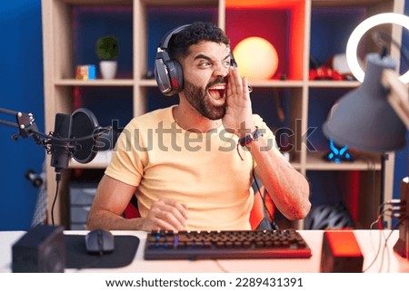 Hispanic man with beard playing video games with headphones shouting and screaming loud to side with hand on mouth. communication concept.  Royalty-Free Stock Photo #2289431391