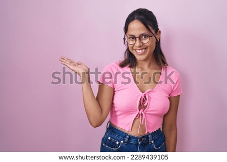 Hispanic young woman standing over pink background wearing glasses smiling cheerful presenting and pointing with palm of hand looking at the camera. 