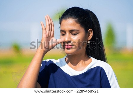 Close up head shot of Indian young woman doing nostril breathing or pranayama yoga or exercise with eyes closed at park - concept of calmness, relaxation, and rejuvenation Royalty-Free Stock Photo #2289468921