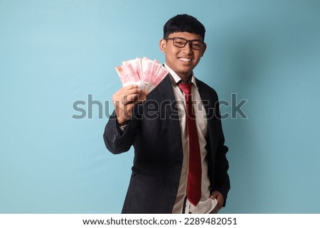 Portrait of young Asian business man in casual suit smiling while holding thousand rupiahs proudly. Isolated image on blue background