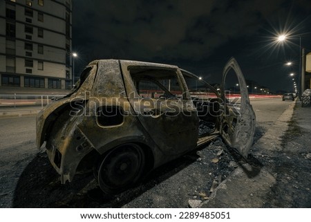 Street view of Milan with a burned down car in the middle of the street, car trails in the night are visible in the background.