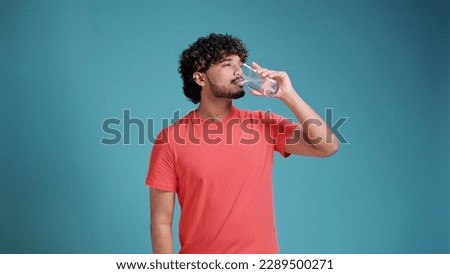 Indian man drinks water from a glass in coral t-shirt on blue studio background. Royalty-Free Stock Photo #2289500271